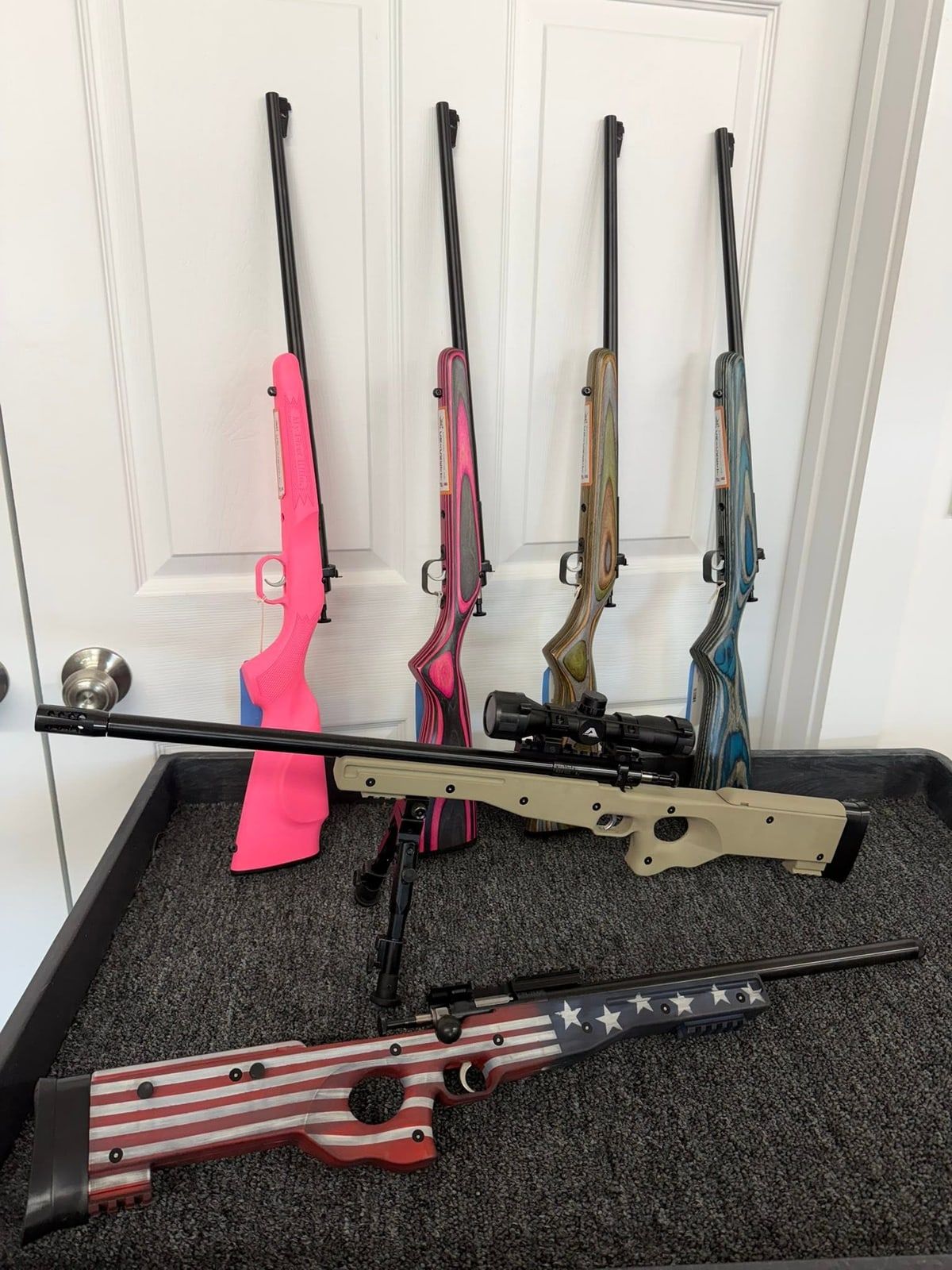 Youth rifles with pink, laminate, and blue stocks, a tan scoped rifle on a bipod, and a flag-painted rifle at New Georgia Pawn